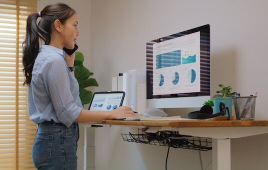 Ergonomischer Homeoffice-Arbeitsplatz mit Computer, Tablet und Telefon, hellem Tageslicht und Pflanzen.