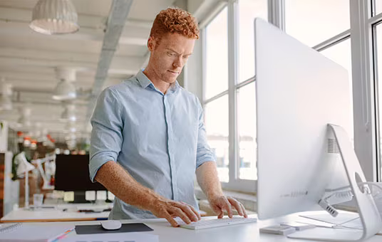 Mann steht am höhenverstellbaren Schreibtisch und arbeitet konzentriert am PC